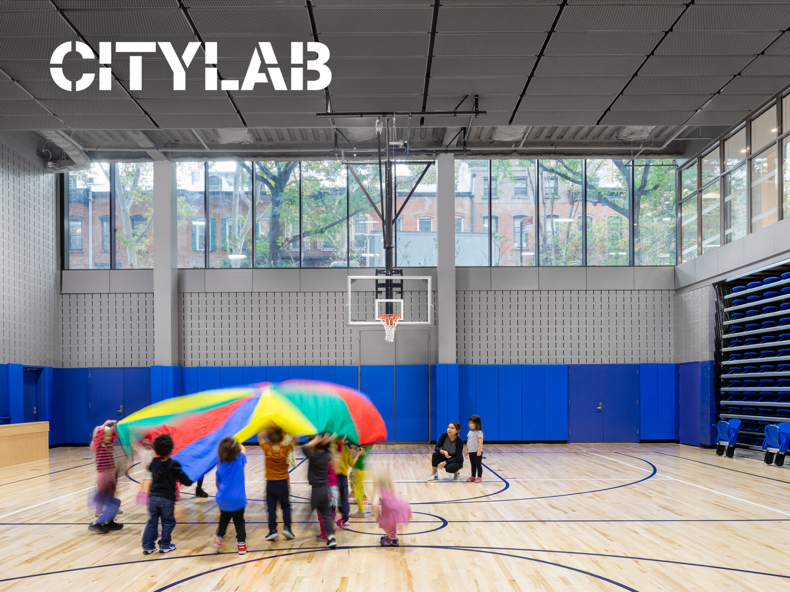 Interior photo of gymnatorium with elementary students playing with rainbow parachute. Photo by James Ewing / JBSA. White logo of Bloomberg CityLab in upper left corner.